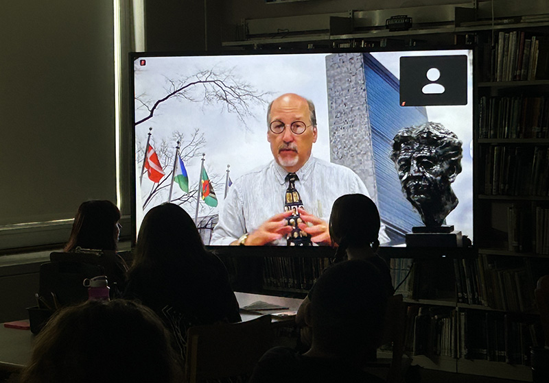 In a darkened room, middle school students are sitting watching a man in a white shirt and tie, with a bust of Eleanor Roosevelt  next to him.