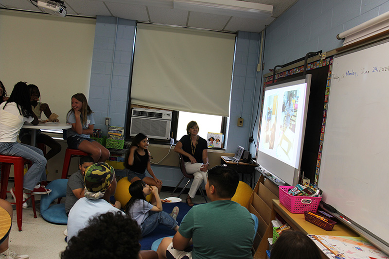 A group of fifth-grade students sit scattered throughout a darkened room. A woman is sitting in the corner. They are all watching a video on a screen at the front of the room.