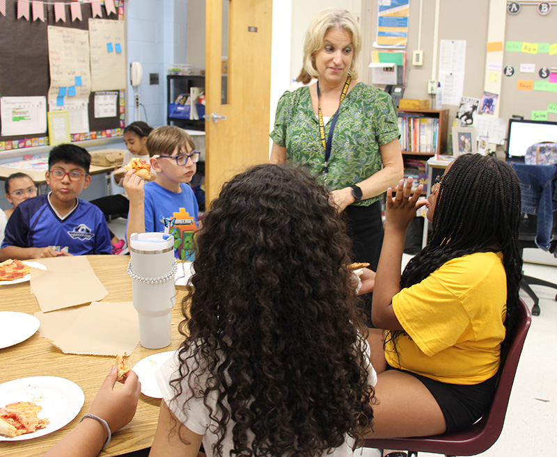 A woman with short blonde hair talks to a couple of fifth-grade girls who are sitting at a table.