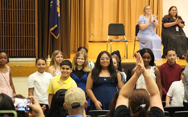A group of fifth-grade students stand in an auditorium facing an audience. Many in the audience are clapping.