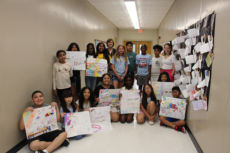 A group of 17 fifth-grade students, some holding posters with art on them, and two women stand together in a hallway, smiling.