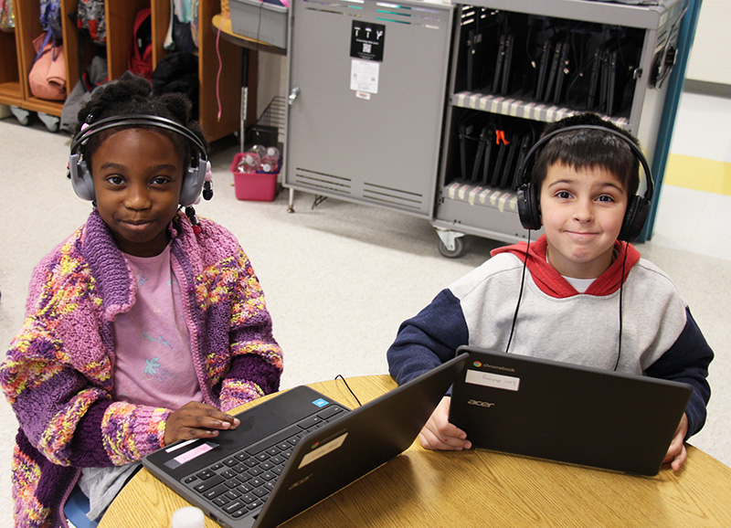 Two first-grade students sit at a table with Chromeboks and wearing headphones. A girl on the left is wearing a pink shirt and a purple print sweater. The boy on the right is wearing a red white and blue sweatshirt and headphones. They are both smiling.