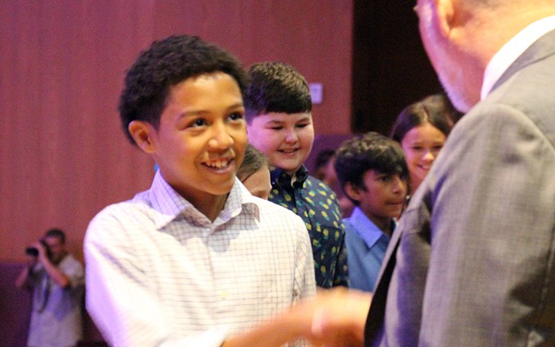 A fifth-grade boy shakes hands with a man. there are many other fifth-graders behind him shaking hands.