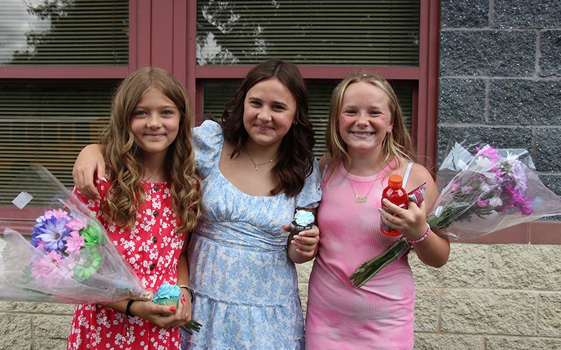 Three fifth-grade girls stand together smiling. They are all wearing nice dresses and holding blue cupcakes and flowers.
