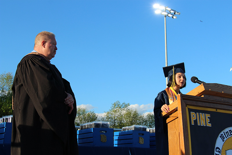 A man in a lack robe stands off to the side as a young woman with short dark hair, wearing a blue cap and gown with a gold sash, speaks at the podium.