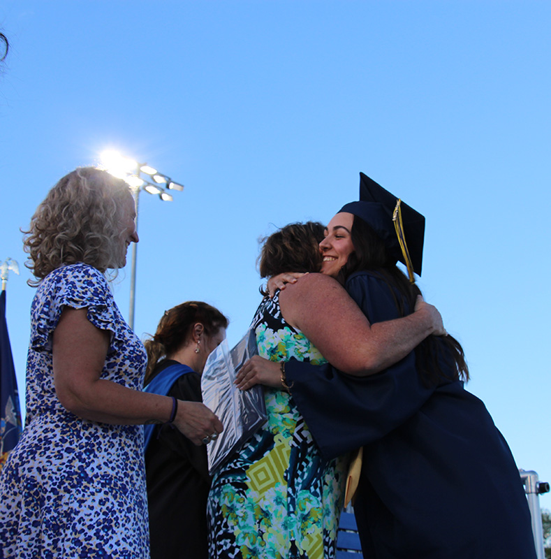 A high school graduate in a blue cap and gown hugs a woman.