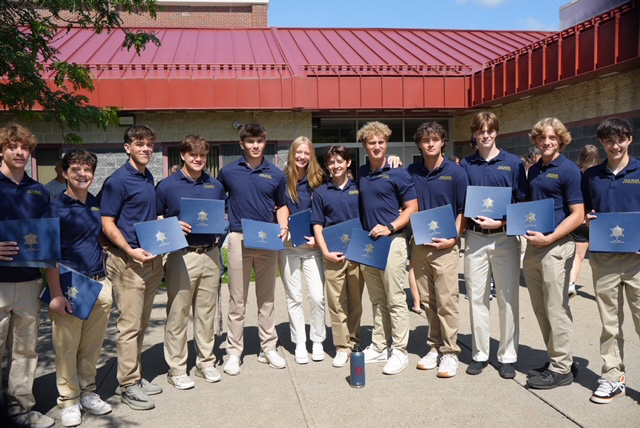 A group of 12 high school students standing together. They are all wearing khaki pants and navy blue shirts. They are holding certificates.