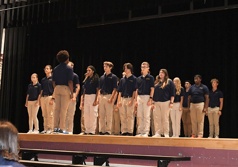 A group of high school students, all wearing khaki pants and blue shirts, line up on a stage.