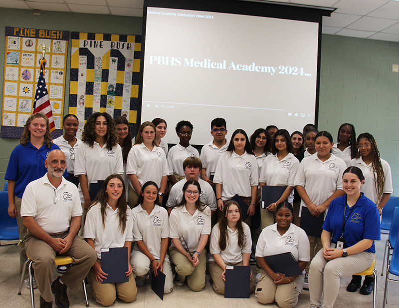 A group of 24 high school students, all wearing white polo shirts, stand or kneel in two rows. There are three adults with them. Behind them is a screen that says PBHS Medical Academy 2024.