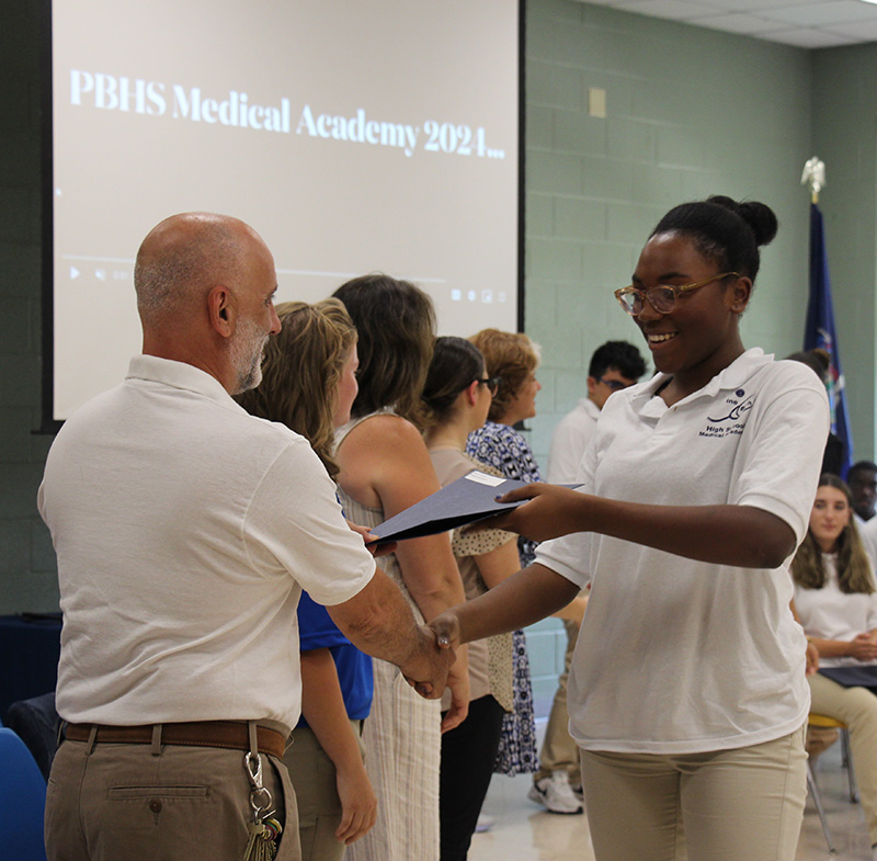 A high school girl with her dark hair up in a bun, accepts a certificate from a man in a white polo shirt.