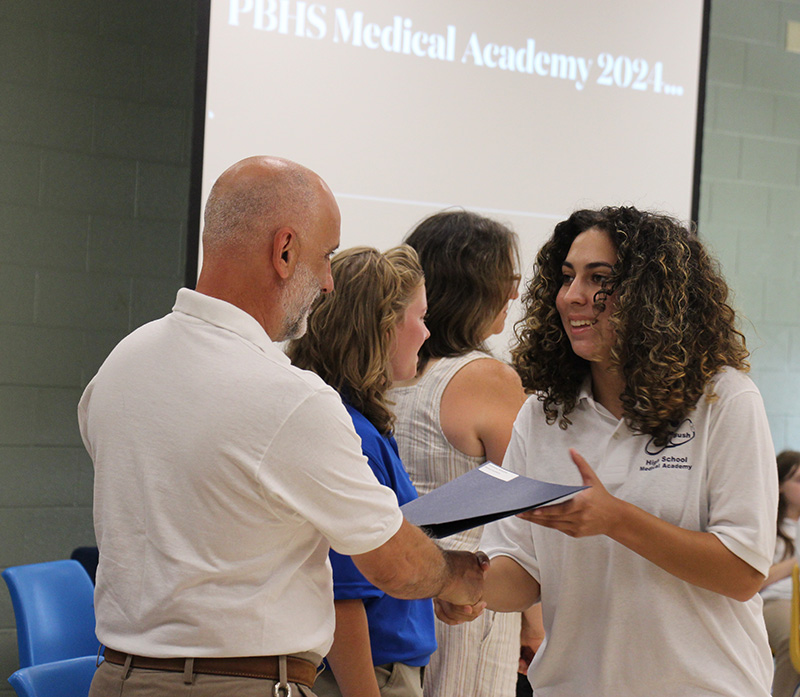 A high school girl with long curly hair, accepts a certificate from a man in a white polo shirt who is shaking her hand.