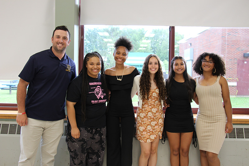 A man stands on the right with five high school young women. They are all smiling.