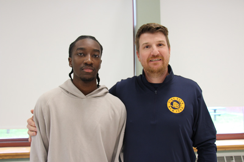 A man on the right and a has his arm around a young man on the left. The young man  is wearing a gray sweatshirt and has dark hair. The man has short light hair and is wearing a navy blue shirt with a gold insignia.