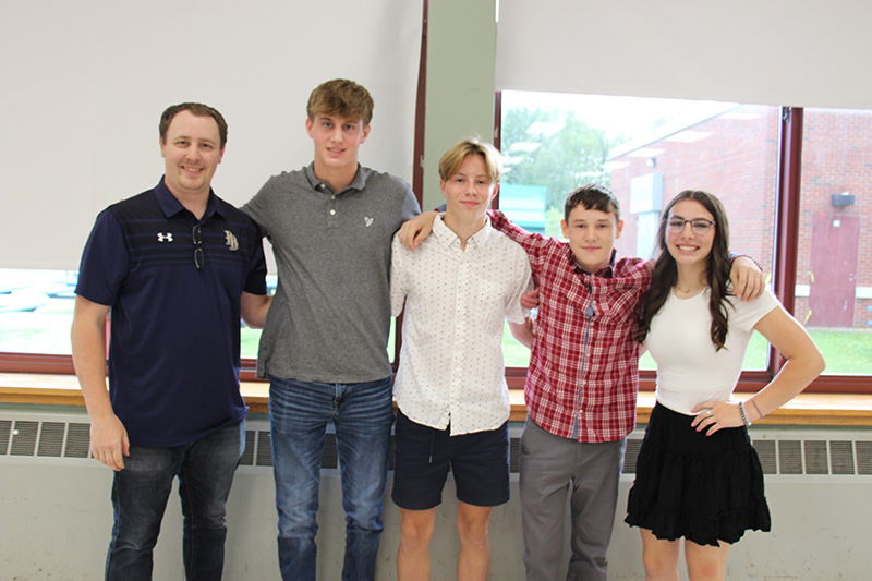 A group of three high school boys and one high school girl stand together with a man on the right. They are all smiling.