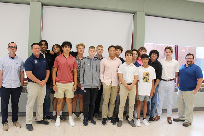 Fifteen high school boys stand in two rows smiling. On the left are two men and on the right is another.