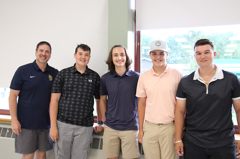 A man wearing a blue polo shirt with the PB logo on the chest pocket stands with four high school boys. They are all smiling.