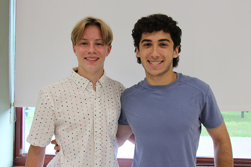 Two high school boy stand together smiling. The kid on the left is wearing a white shirt and has short blonde hair. The kid on the right has dark hair and is wearing a blue shirt.