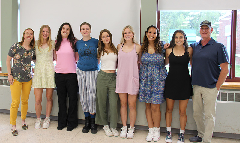 A man stands on the right and a woman on the left. In between them are seven high school young women smiling. They are all arm in arm.