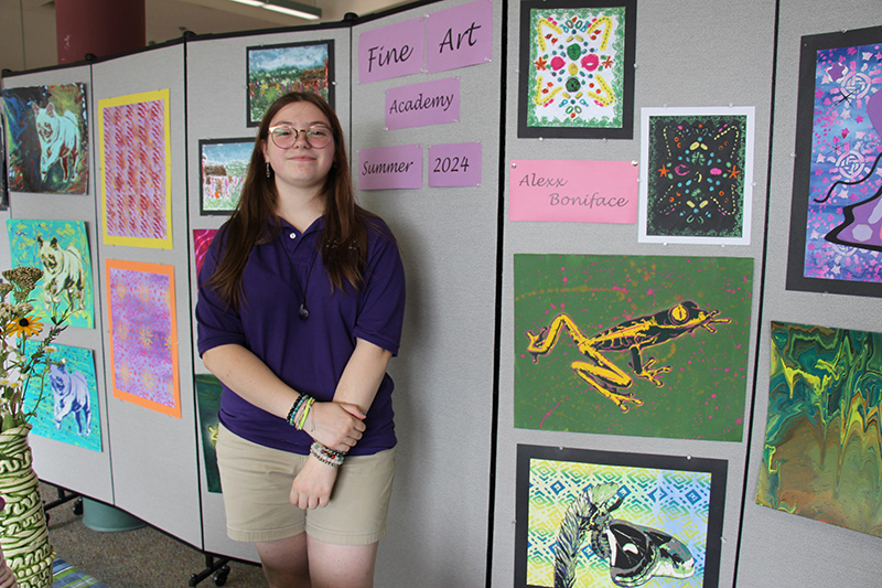 A high school girl with long dark hair, wearing glasses and a purple shirt, stands in front of a wall with her artwork handing.