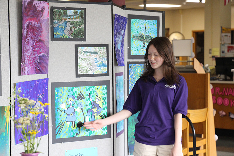 A high school girl wearing a purple shirt with shoulder-length dark hair, points to a painting she made and talks about it.