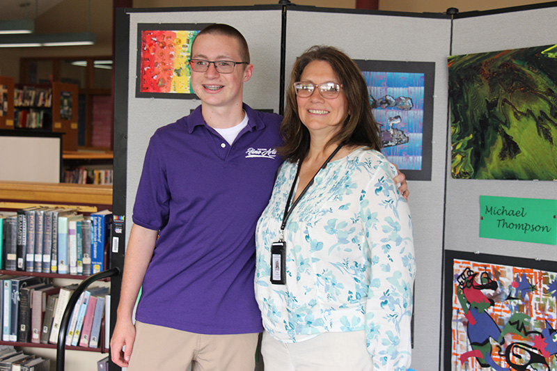 A high school boy with short hair and glasses, wearing a purple polo shirt, stands in front of artwork he made. He has his arm around a woman. Both are smiling.