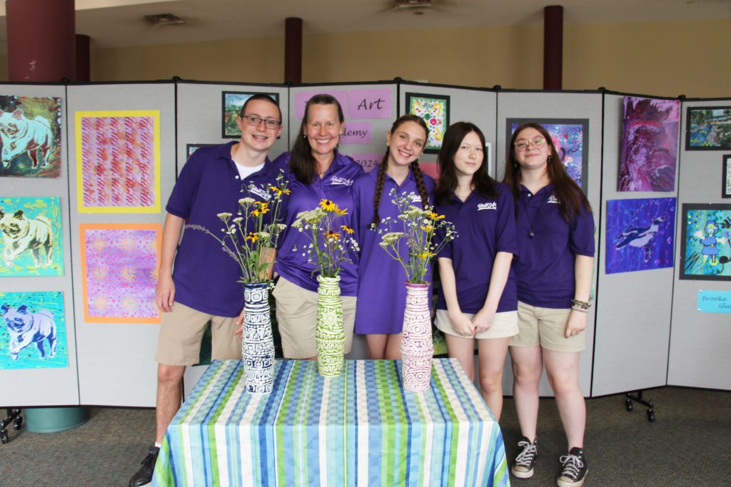 A group of five people, all wearing purple polo shirts, stand around a table that has three tall vases on them. The high school kids made them. All are smiling.