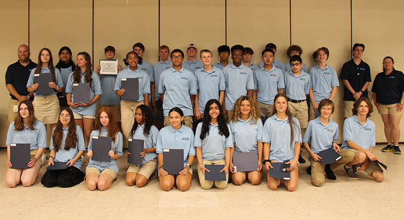 A group of 29 high school students stand in two rows, first row kneeling and second row standing. There are three adults on the ends. They are all wearing light blue polo shirts that say Aviation Academy.