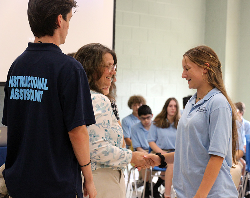 A high school girl with long light hair shakes the hand of a woman with medium length dark hair. They are both smiling.