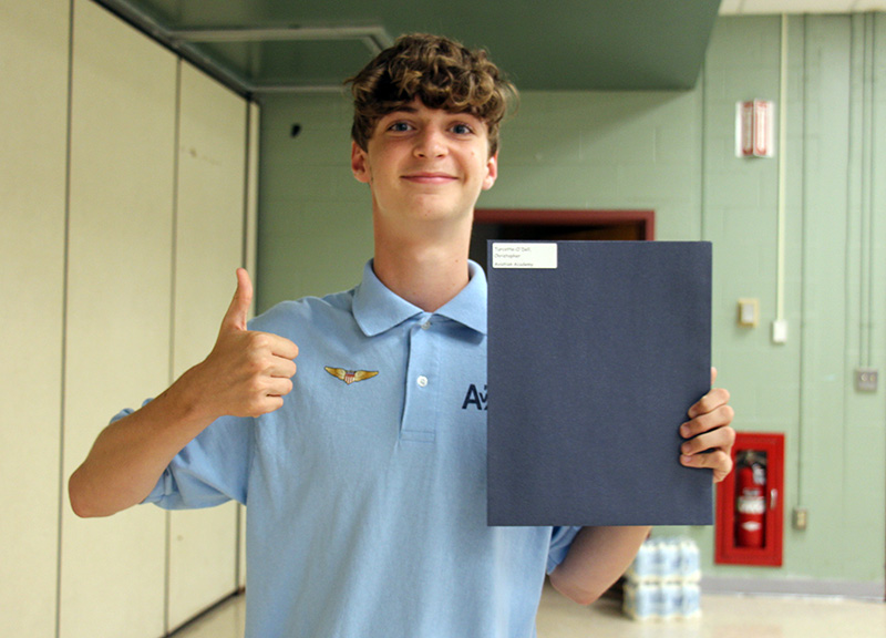 A high school boy with short dark hair wearing a light blue polo shirt gives a thumbs up and holds a blue folder with a certificate in it in his other hand. He is smiling.