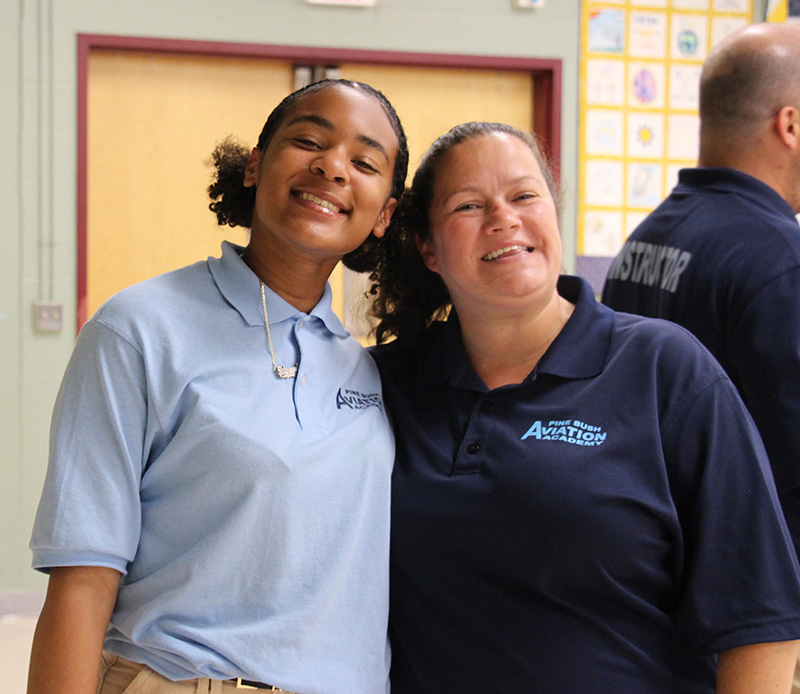 A high school girl, wearing a light blue polo shirt smiles. She has shorter dark ponytails. She is standing with a woman in a dark blue polo shirt who is also smiling.