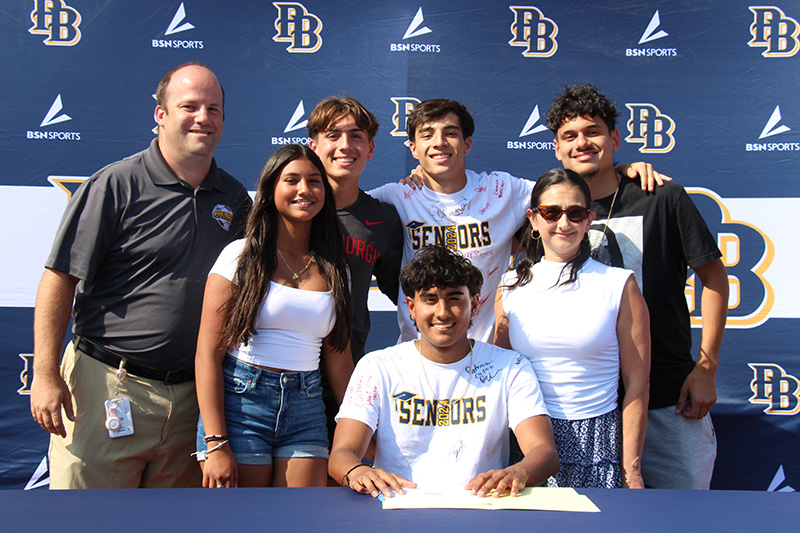 A group of six people - one man and five high school kids - stand behind a young man swho is sitting at a table signing a piece of paper. They are all smiling.