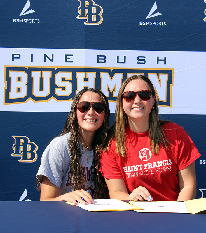 Two high school girls sit together smiling.  They both have long hair and sunglasses on.  Behind them is a sign that says Pine Bush Bushmen.