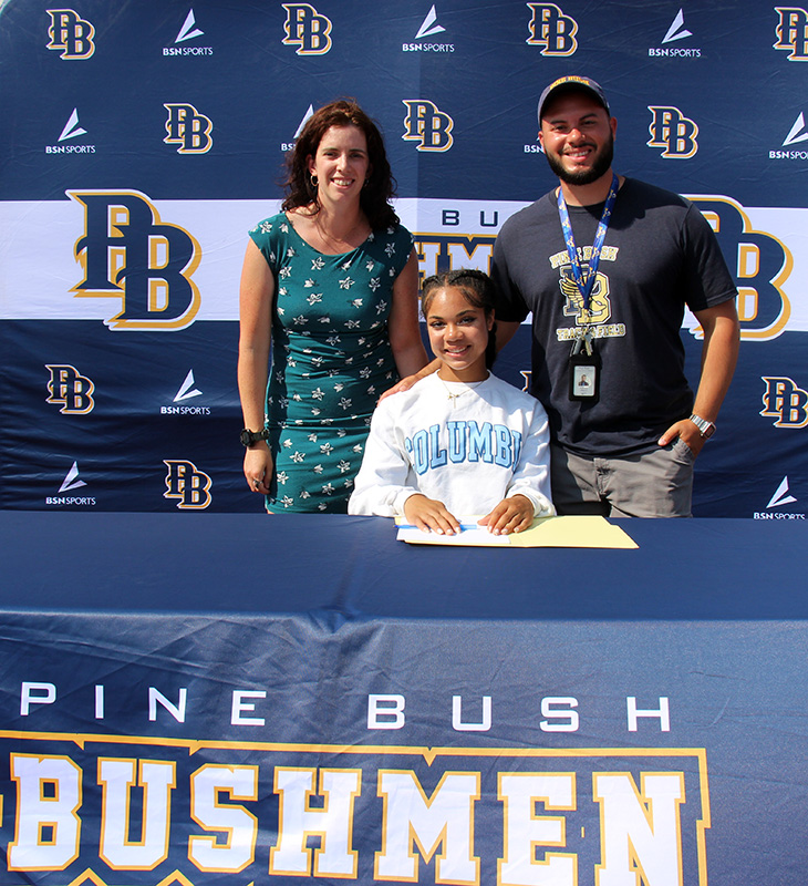 A man and woman stand behind a high school girl who is sitting at a table signing a paper.  The tablecloth says Pine Bush Bushmen.  they are all smiling.