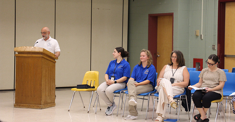 A man in a white polo shirt stands at a podium speaking as four women sit in chairs next to him listening.