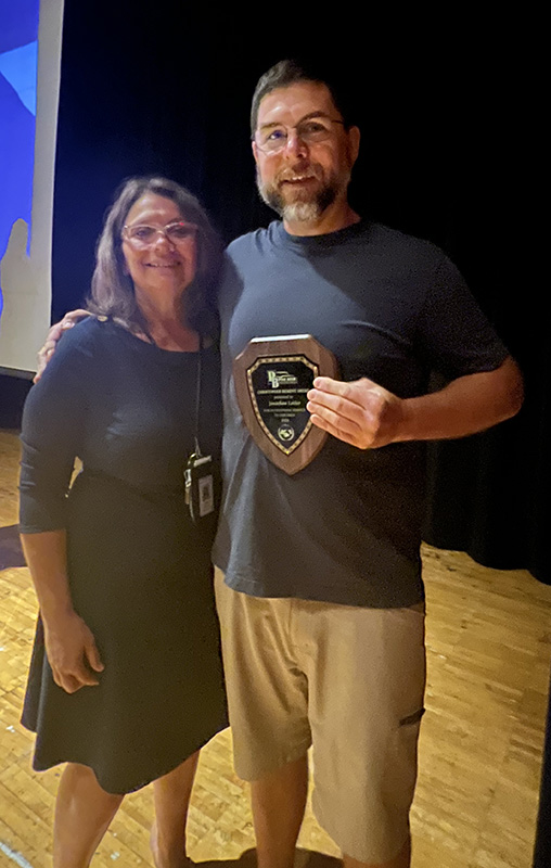 A man on the right with short dark hair and a beard, wearing a blue shirt and tan shorts, holds a plaque. He is standing on a stage with a woman in a dark blue dress, with longer dark hair. they are both smiling.