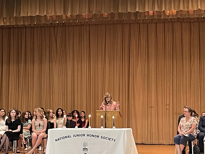 a woman stands at a podium speaking. Behind her are middle school students sitting in chairs on a stage. In front of her is a table with four candles on it. The tablecloth says National Junior Honor Society.