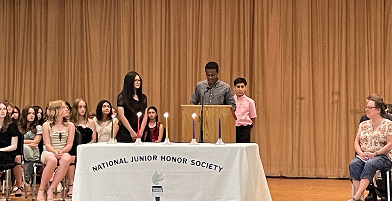 Three middle school students stand around a table with four candles on it. The tablecloth says National Junior Honor Society. One student is at the podium speaking.