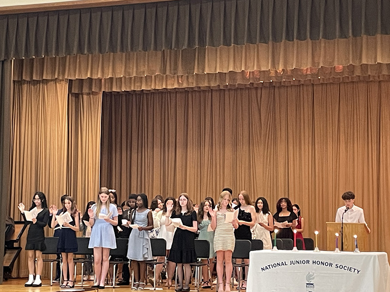 A group of about 20 middle school students stand on a stage, all dressed nicely. They have  their hands up pledging to the national junior honor society.