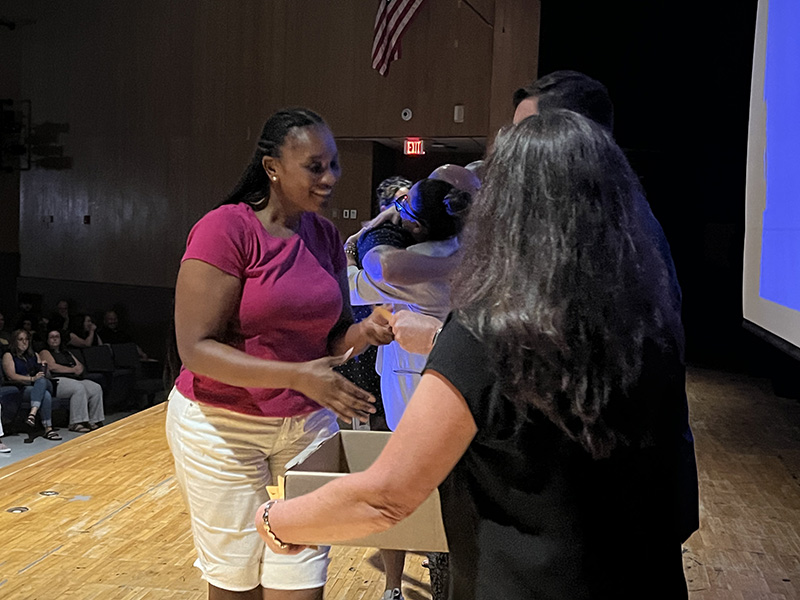 A woman with a pink shirt and white pants shakes hands with someone on a stage.