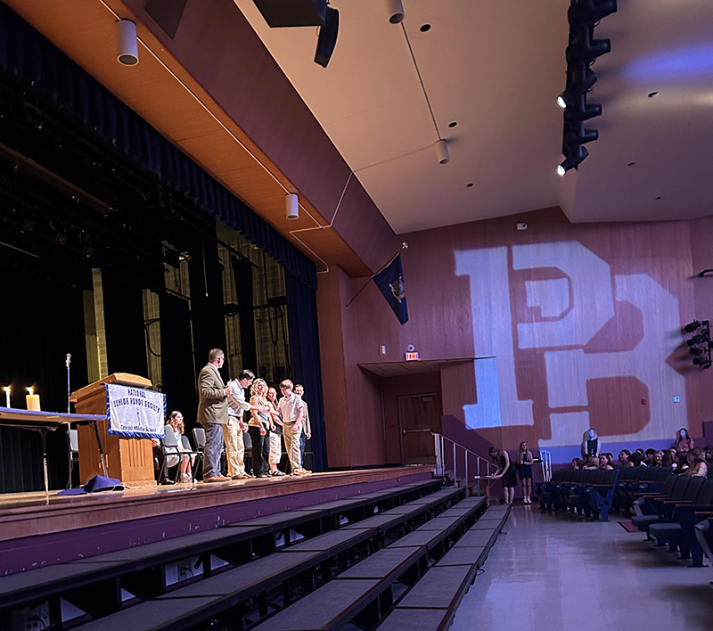 A view of an auditorium from the side. There are people and a podium on the far side and a large PB reflected on the wall.