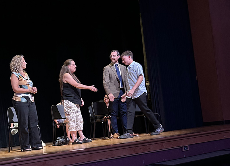 A middle school student shakes hands with adults on a stage.