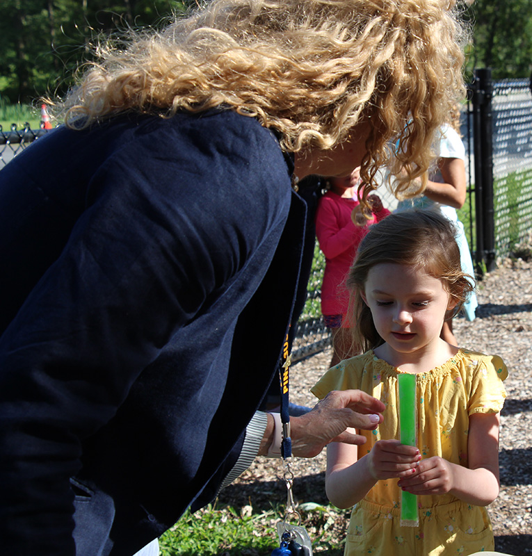 A woman with long blond hair, wearing a blue jacket, offers a green frozen popsicle to a little girl wearing a yellow shirt.