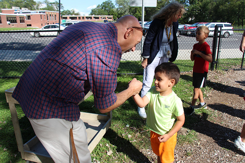 A man with a blue and red checked shirt gives a fist bump to a little boy with dark hair, wearing orange shorts and a green shirt.