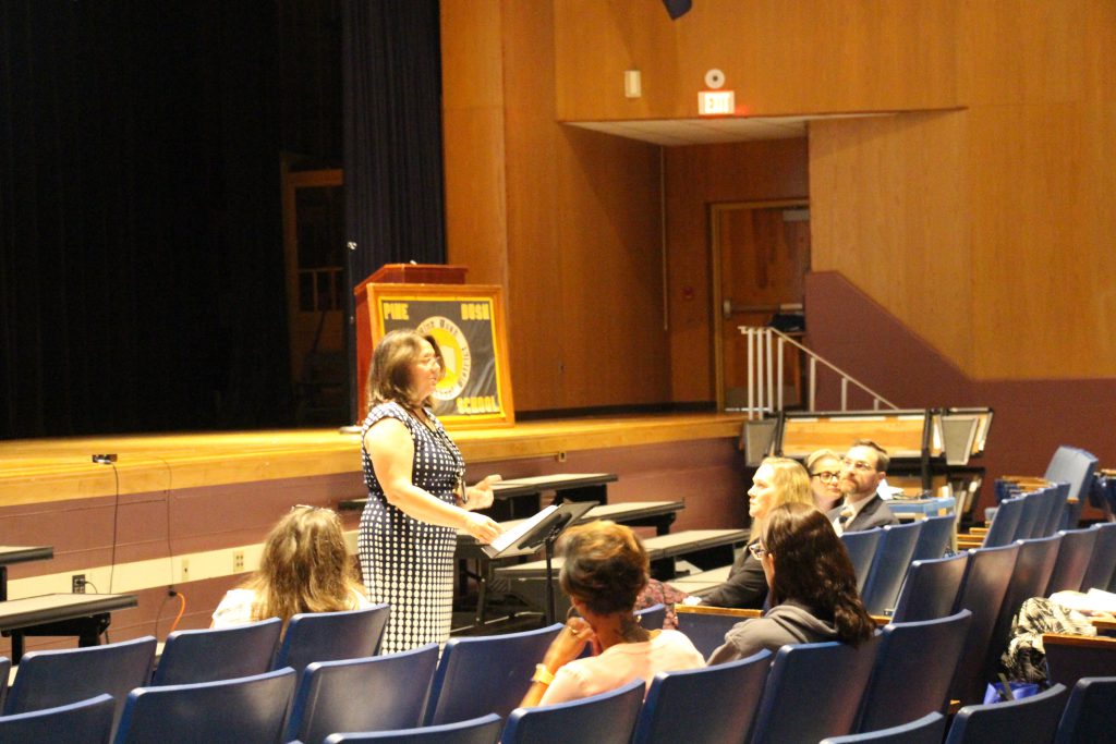 A woman in a blue and white printed dress talks to people in an auditorium.