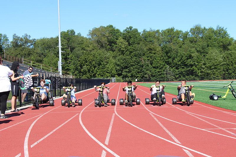 A clay colored track with six high school students each sitting on a low to the ground trike, ready to race.