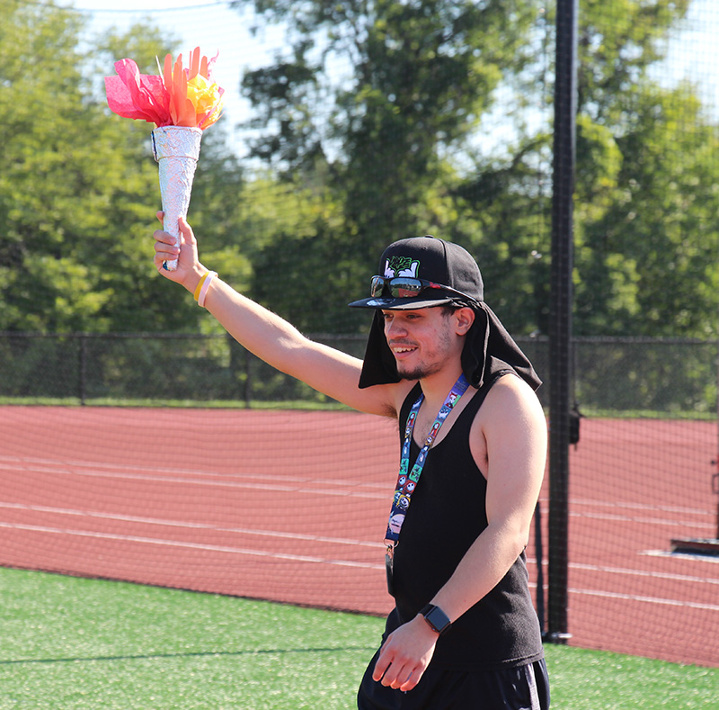 A high school boy, dressed in black shirt and shorts with a black baseball hat on holds up what looks like an olympic torch made of paper.