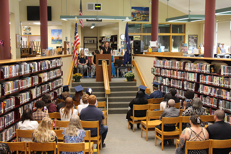 Taken from the back of the room, people sit on both sides of an aisle leading up to a set of stairs. At the top of the stairs are more adults standing.