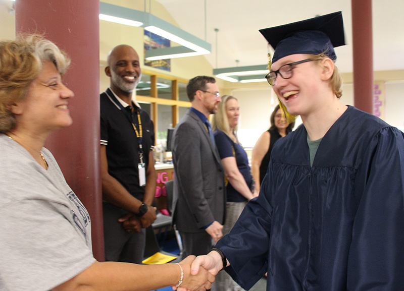 A high school senior, with blonde hair, wearing glasses and a blue cap and gown, shakes a woman's hand. They are both smiling.