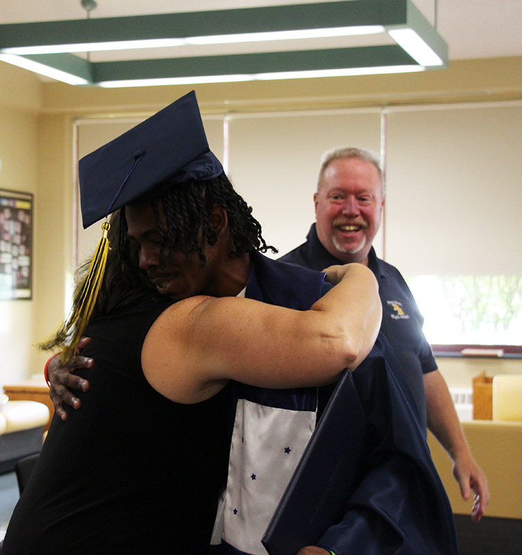 A high school student wearing a navy blue cap and gown hugs a woman. A man standinggbehind them smiles.