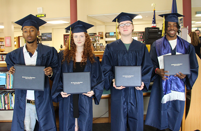 Four high school seniors all wearing navy blue caps and gowns stand next to each other holding their diplomas. There are three boys and one girl. Some are smiling.